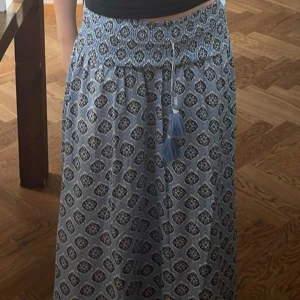 Blåvit mönstrad långkjol med tofsar - Säljer en blåvit långkjol med elastisk midja och dekorativa tofsar. Kjolen har ett geometriskt mönster och är luftig och skön, perfekt för varma dagar. Passar till både strand och stad. Är i storlek 164 men passar en xs