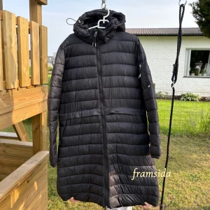 Svart lång dunjacka från CRW - ( ridjacka ) Säljer en svart lång dunjacka från CRW med huva och dragkedja framtill. Jackan har quiltad design och är perfekt för kyliga dagar. Passar dig som vill ha en stilren och varm jacka med praktiska detaljer. Går att knäppa upp baktil så man kan rida/vara rörlig. 