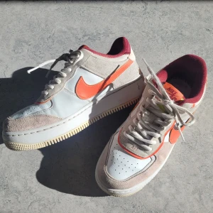 Nike Air Force 1 sneakers med orange detaljer - Nike Air Force 1 sneakers i vitt och beige skinn och mocka, med orange swoosh och detaljer. Insidan är vinröd och sulan är vit med beige undersida. Klassisk modell med snörning och AIR-text i gult på hälen. Skorna är i god kondition och har inga skador. 