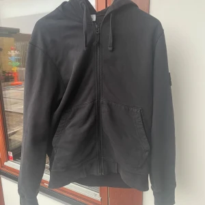 Svart hoodiejacka från Stone Island - Säljer en svart hoodiejacka från Stone Island med dragkedja framtill och huva med snörning. Jackan har två fickor på magen och klassisk Stone Island-logga på ärmen. Perfekt för en avslappnad och stilren look. Jag har använt hoodien en del men väldigt sparsamt och tänker att 1200 är rimligt pris! Självklart äkta som är köpt på NK, kvitto ingår!