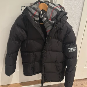 Svart dunväst från Burberry - Snygg svart dunväst från Burberry med avtagbar huva och dragkedja framtill. Västen har två fickor med dragkedja och en klassisk Burberry-logga på vänster ärm. Fodret har ett grått rutigt mönster med röda detaljer. Storlek L/M