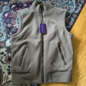 Ralph Lauren purple label vest  - Stilren beige väst från Ralph Lauren med hög krage och dragkedja framtill. Västen har två fickor med dragkedja och diskret broderad logga på bröstet. Ena sidan är i mjuk bomull, andra i slät syntet för en sportig look.