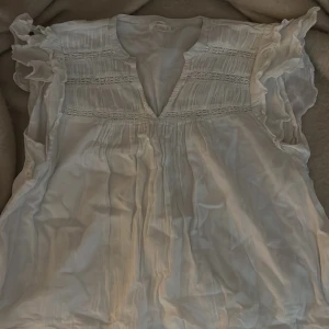 Vit blus med volang från Gina Tricot - Säljer en söt vit blus från Gina Tricot med volangdetaljer på ärmarna och v-ringning. Blusen har fina spetsinslag och veck framtill som ger en romantisk känsla. Perfekt till vår och sommar! Använd max två gånger!
