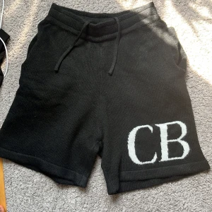 Svarta stickade shorts från Cole Buxton - Svarta stickade shorts från Cole Buxton med vita initialer 'CB' på vänster ben. Shortsen har dragsko i midjan och två bakfickor. Perfekta för en avslappnad och trendig stil. Pris kan diskuteras 