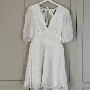 Vit omlottklänning från Gina Tricot - Superfin vit omlottklänning från Gina Tricot med v-ringning, puffärmar och knyt i ryggen, skyddande underkjol under så den är inte genomskinlig. Klänningen har en markerad midja och är tillverkad i ett lätt, plisserat material som ger en luftig känsla. Perfekt för sommarens alla tillfällen eller student! Använd endast en gång och har inga tecken på användning.