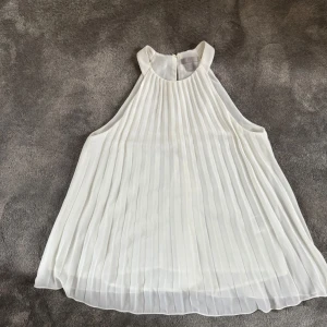 Vit plisserad ärmlös blus - Superfin vit blus från H&M med plisserat tyg och hög hals. Blusen är ärmlös och har en luftig, A-linjeformad passform som ger ett elegant intryck. Perfekt för sommarens tillfällen och enkel att styla med accessoarer.