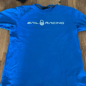 Blå t-shirt från Sail Racing - Säljer en blå t-shirt från Sail Racing med vit logga tryckt på bröstet och en liten logga i nacken. Klassisk passform och rund halsringning. Perfekt för dig som gillar stilren design och sportiga märken.