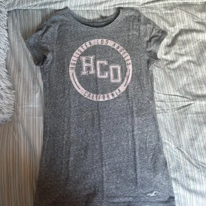 Grå t-shirt från Hollister - Grå t-shirt från Hollister med tryck 'HCO Hollister Los Angeles California' i rosa framtill. Klassisk rund halsringning och korta ärmar. Oanvänd. 