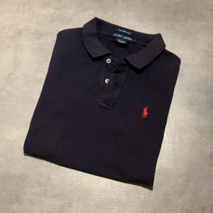 Mörkblå pikétröja från Ralph Lauren - Mörkblå pikétröja från Ralph Lauren med klassisk krage och knappar framtill. Röd broderad logga på bröstet, RL13 broderat nertill och TEAM PESTANA broderat på ryggen. Perfekt för en sportig och stilren look. Tröjan är i storlek L för barn. Bara att höra sv sig vid frågor eller funderingar!