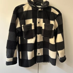 Svartvit rutig teddyjacka från H&M - Mysig teddyjacka från H&M i svartvitt rutigt mönster. Jackan har två stora bröstfickor, knappar framtill och en klassisk krage. Perfekt för kyliga dagar när du vill vara både varm och snygg. Storlek 140 cm, 8-10 år