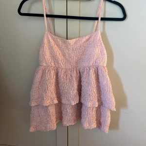 Ljusrosa volangtop  - Supersöt ljusrosa topp från Gina med smala axelband och volanger i flera lager. Toppen har en skrynklig struktur och är perfekt för varma dagar. Passar till både jeans och kjol för en söt look.