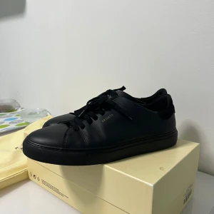 Svarta sneakers från Axel Arigato - Stilrena svarta sneakers från Axel Arigato med diskret logga i guld på sidan. Skorna har snörning, rund tå och är tillverkade i skinn. Perfekta för en clean och modern look. Strl 44.5 helt nya