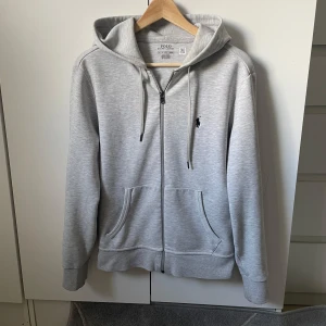 Polo zip hoodie - Säljer en stilren ljusgrå hoodie från Polo Ralph Lauren i storlek XS. Tröjan har dragkedja, huva med snörning och två fickor framtill. Klassisk broderad logga på bröstet. Perfekt för en avslappnad look.