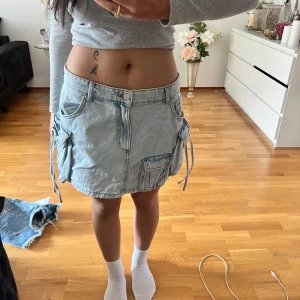 Ljusblå jeanskjol med fickor och snörning - Trendig ljusblå jeanskjol med stora cargofickor på sidorna och cool snörningsdetalj. Kjolen har normal passform och klassisk knapp- och dragkedjestängning. Perfekt för dig som gillar streetstyle och vill sticka ut.