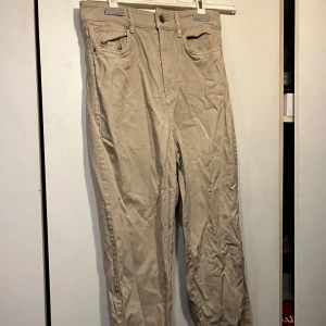 Beige byxor från H&M - Säljer ett par klassiska beige byxor från H&M med normal passform. Byxorna har bälteshällor och stängs med dragkedja och knapp. Perfekta till vardags och enkla att matcha med olika toppar.
