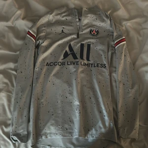 PSG x Jordan långärmad fotbollströja - Snygg långärmad PSG fotbollströja i grått med svarta prickar, Jordan-logga och Paris Saint-Germain-märke på bröstet. Röd, vit och svart detalj på ärmarna. Dragkedja vid halsen och tryck på både fram- och baksida.