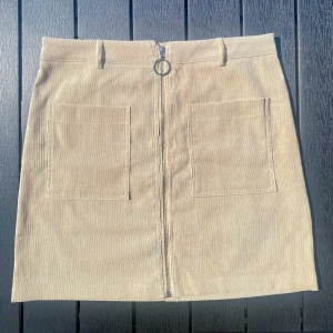 Beige kjol i manchestertyg - Säljer en beige kjol i manchestertyg med två stora fickor framtill, en silverfärgad dragkedja och hällor för bälte. Säljer den pga av jag tycker den är lite för kort på mig som är 180. 