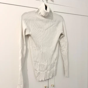Vit ribbad polotröja - Säljer en stilren vit polotröja med ribbad struktur. Tröjan har lång ärm och hög polokrage, perfekt för lager-på-lager. Passar till många olika outfits och är ett måste i garderoben.