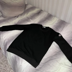 Svart sweatshirt från Moncler - Säljer en svart sweatshirt från Moncler med diskret logga i samma färg på bröstet och Moncler-märke på ena ärmen. Tröjan har rund halsringning och långa ärmar. Perfekt för en stilren och enkel look. Passar även S
