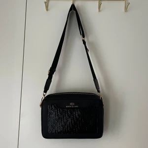 Svart axelremsväska från River Island - Snygg svart axelremsväska från River Island med bred justerbar axelrem och guldfärgade detaljer. Väskan har en glansig frontficka med präglat mönster och en dekorativ R-berlock i metall. Perfekt för att bära dina viktigaste saker med stil. Har används ett få antal gånger. Priset kan diskuteras. 