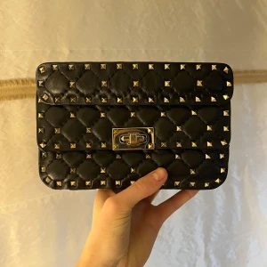 Svart axelväska med nitar från Valentino - Säljer en svart quiltad axelväska från Valentino med silverfärgade nitar och vridlås. Väskan har ett handtag upptill och insida i rött. Perfekt för dig som vill ha en edgy accessoar med lyxig känsla.