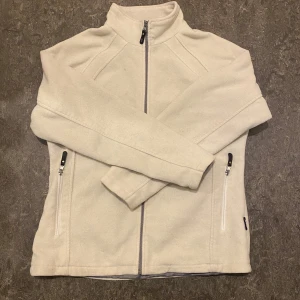 Vit fleecejacka från True North - Mysig vit fleecejacka från True North med hög krage och hel dragkedja framtill. Jackan har två sidofickor med dragkedja och en enkel, stilren design. Perfekt för lager-på-lager under kyliga dagar. Storlek L, men passar som M