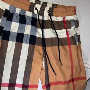 Rutig shorts från Burberry London - Snygga shorts från Burberry London med klassiskt rutigt mönster i beige, brunt, svart, vitt och rött. De har elastisk midja med svart snörning och metalländar. Perfekta för sommaren och har en modern passform. (Storlek M men passar S också kartong på)