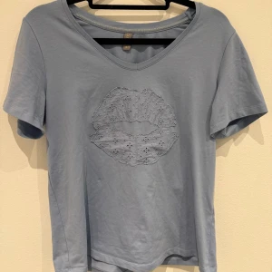 Ljusblå t-shirt med spetsdetalj - Säljer en ljusblå t-shirt med v-ringning och korta ärmar. På bröstet finns en stor applikation i form av läppar gjord av broderad spets, vilket ger en unik och trendig look.