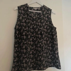 Svart blommig ärmlös blus - Säljer en svart ärmlös blus med små blommor i ljusrosa och vitt. Blusen har en lätt v-ringning och ett luftigt fall, perfekt för varmare dagar. Passar till både jeans och kjol.