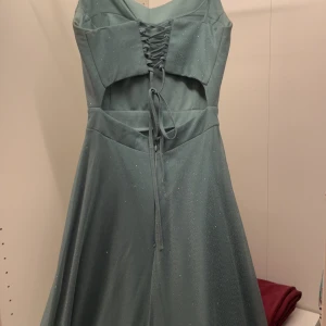 Glittrig grön/blå  bal/bröllop/fest långklänning med snörning, corset rygg. - grön långklänning med glittrigt tyg och smala axelband. Klänningen har snörning framtill, cut-out-detalj. Midja ungefär 64 cm. Från midja-nedåt: 107 cm. Köpt utomlands. OBS! Slitage på änden pga klippning (förkortning)