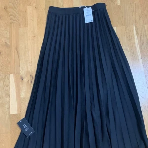 Svart plisserad långkjol från NA-KD - Säljer en svart plisserad långkjol från NA-KD. Kjolen har klassiska veck och en hög midja som ger en snygg siluett. Perfekt att styla med en enkel topp eller stickad tröja.