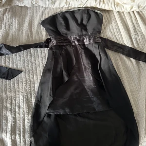 Svart  långklänning med pärldetalj - Säljer en svart axelbandslös långklänning med pärldetalj längs överkanten. Klänningen har omlottliknande lager framtill och ett transparent, längre tygparti baktill. Sidenliknande material och markerad midja med band som kan knytas. Den är lite smutsig vid den transparenta längre delen, därav billigare pris.