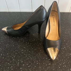 Svarta pumps med gulddetalj från SthlmDG - Snygga svarta pumps från SthlmDG med spetsig tå och guldfärgad metall-detalj framtill. Skorna har hög klack och är tillverkade i ett slätt material med beige innersula. Perfekta för att lyfta din outfit.