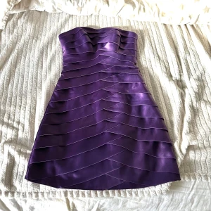 Lila kortklänning - Säljer en snygg lila axelbandslös kortklänning med vågformade banddetaljer över hela klänningen. Klänningen har dragkedja i sidan och är figurnära i modellen. Perfekt för dig som vill sticka ut med en färgstark look.
