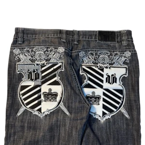 Gråa jeansbyxor med broderad sköld på bakfickorna - Säljer ett par gråa jeansbyxor från Victorious med stora broderade sköldar och kronor i svart och vitt på bakfickorna. Byxorna har vida ben och klassisk femficksdesign. Perfekta för dig som vill sticka ut med detaljerad brodyr. Grå/svarta