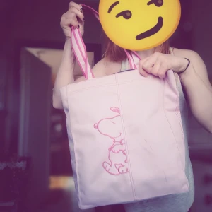 Rosa shoppingväska med Snoopy-tryck - Säljer en söt rosa shoppingväska med tryck av Snoopy på framsidan. Väskan har två handtag i matchande färg och en praktisk dragkedjeficka på baksidan. Perfekt för att bära med sig böcker, laptop eller annat smått och gott.