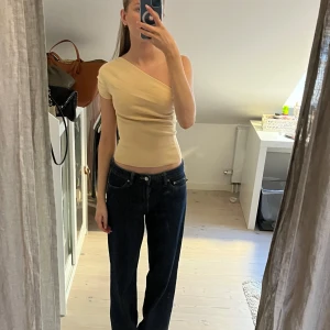 Beige one shoulder topp - Snygg beige topp med one shoulder-design och tight passform. Toppen har en stilren look och är perfekt för att lyfta en outfit med sin asymmetriska skärning.