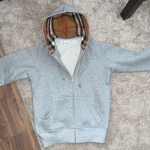 Burberry Hoodie - Säljer en grå hoodie från Burberry med dragkedja och två fickor framtill. Huvan är fodrad med Burberrys ikoniska beige, röda och svarta rutmönster. Perfekt för en stilren look. Bara att höra av sig vid eventuella frågor eller funderingar.