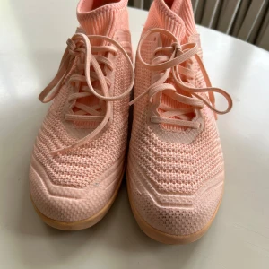 Adidas Predator rosa fotbollsskor - Säljer ett par ljusrosa Adidas Predator fotbollsskor med stickad ovandel och snörning. Skorna har gummisula med mönster för bra grepp och klassiska Adidas-ränder på sidan. Perfekta för inomhusspel och har en modern, sportig look.