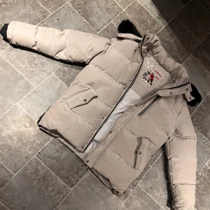 Beige dunjacka från Moose Knuckles - Säljer en beige dunjacka från Moose Knuckles med svart pälskant på huvan som kan tas bort om man vill. Guld detaljer och MK loggan i guldfärg. Jackan har dragkedja, stora fickor och broderad hockeypatch på insidan. Inga skador på jackan. Mvh 