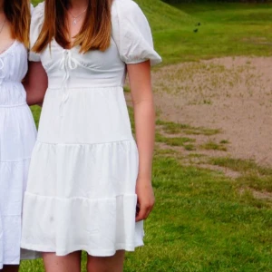 Vit kortklänning med puffärm från Gina Tricot - Supersöt vit miniklänning från Gina Tricot med puffärmar och knytning vid bysten. Klänningen har volanger i kjolen och en lätt rynkad struktur i tyget. Perfekt för sommarens alla tillfällen!