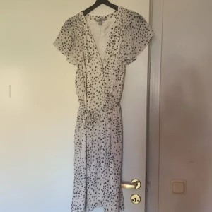 Vit blommig klänning från H&M - Söt vit klänning från H&M med små svarta blommor, v-ringning och knytband i midjan. Klänningen har korta, luftiga ärmar och är tillverkad i ett lätt och skönt material. Perfekt för varma dagar! Storleken är xl men passar bra på m också