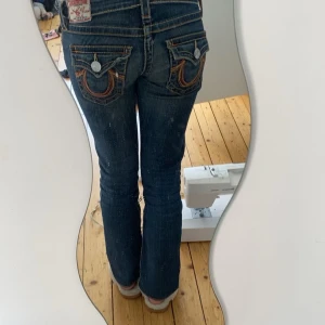 Mörkblå jeansbyxor från True Religion - Säljer ett par mörkblå jeans från True Religion med kontrastsömmar och dekorativa fickor bak. Byxorna har låg midja, bootcut-modell och klassiska detaljer som knappar och dragkedja framtill. Säljer på grund av att dem är kanske 3-5 cm för korta på mig skulle säga att dem skulle passa någon i ungefär 160 längden. Storlek W25 skriv om mer frågor!!