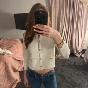 Vit kofta från Zara med guldknappar - Säljer en stilren vit kofta från Zara med dekorativa guldknappar framtill och två små fickor. Koftan har rund halsringning och långa ärmar. Perfekt för lager-på-lager och enkel att matcha med olika outfits.