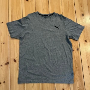 Grå Puma t-shirt - Säljer en grå t-shirt från Puma med klassisk passform och svart Puma-logga på bröstet. T-shirten är tillverkad i mjuk bomull och har korta ärmar. Perfekt för en avslappnad stil.
