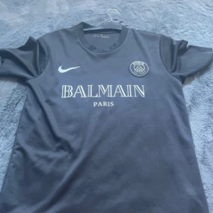 Svart PSG fotbollströja Nike x Balmain - Säljer en svart PSG fotbollströja från Nike med Balmain Paris-tryck på bröstet. Tröjan har korta ärmar, rund hals och är tillverkad i Dri-FIT-material för bra andningsförmåga. PSG-logga och Nike swoosh på framsidan.