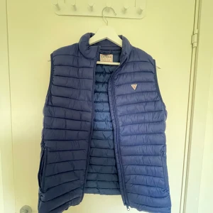 Blå dunväst från Guess - Snygg blå dunväst från Guess med quiltad design och dragkedja framtill. Västen har ståkrage och två fickor med dragkedja på sidorna. Perfekt för lager-på-lager och enkel att matcha med olika outfits.