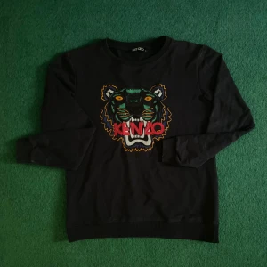 Svart sweatshirt med tigerbrodyr från Kenzo - Mycket fint skick, knapen använd. Inga defekter!Svart sweatshirt från Kenzo med ikonisk tigerbrodyr och färgglada detaljer framtill. Tröjan har rund halsringning och långa ärmar. Perfekt statement-plagg för dig som gillar streetwear och snygga loggor.