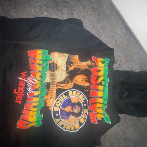 Svart hoodie med Bob Marley-tryck från Wrangler - Säljer en svart hoodie från Wrangler med stort färgglatt Bob Marley-tryck och texten 'Positive Vibrations' på ryggen. Framtill finns en liten Wrangler-logga i rött. Tröjan har huva och dragsko.