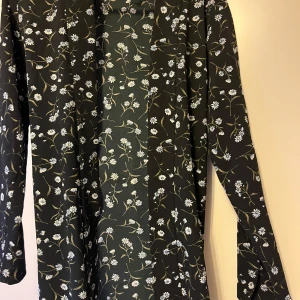 Svart blommig klänning från Divided - Svart mönster blus med band  från Divided med små vita och blå blommor samt gröna blad. Blusen  har lång ärm och ett knytband i midjan för en smickrande passform. Perfekt för dig som gillar blommiga mönster och vill ha något bekvämt och snyggt.